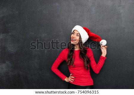 Happy brunette woman in red blouse and christmas hat holding arm on hip and looking away over black background
