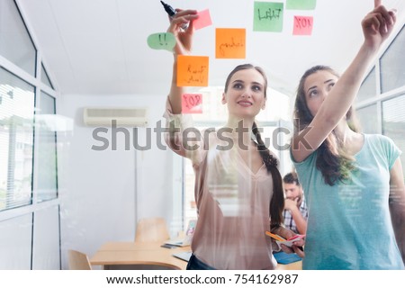 Two proficient female co-workers posting sticky notes in the interior of a shared office space for task prioritization and better time management Royalty-Free Stock Photo #754162987