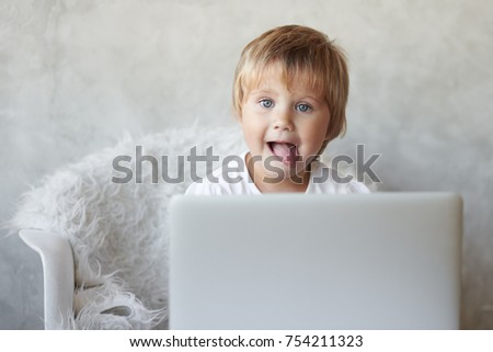 Childhood, modern technology, electronic gadgets and addiction concept. Funny Caucasian little girl with short hairstyle using generic notebook pc, watching cartoons online, sticking out tongue