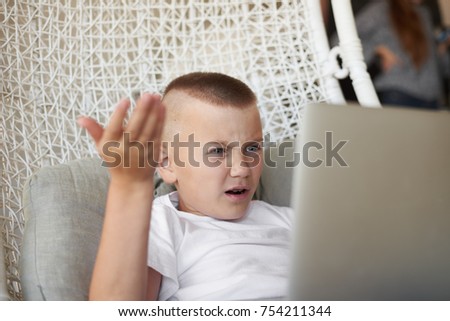 Indoor picture of angry displeased school boy sitting on woven armchair with generic portable computer and gesturing in indignation, feeling annoyed and furious while losing video game again