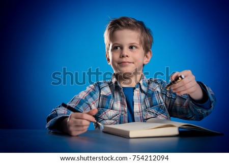 Cute little boy schoolboy in checkered shirt sits at a table and writes in a notebook on a blue background