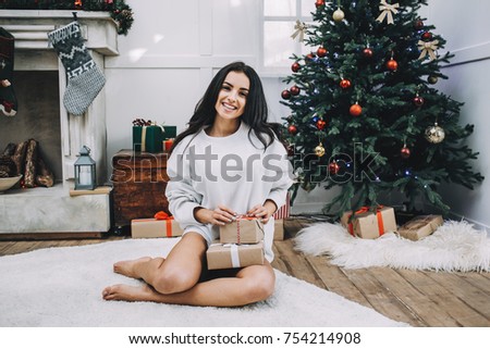 Another picture of attractive girl sitting on the white cozy carpet and smiling to the camera. She holds presents for her family on her knees and waiting for them in the room. Behind her there are a