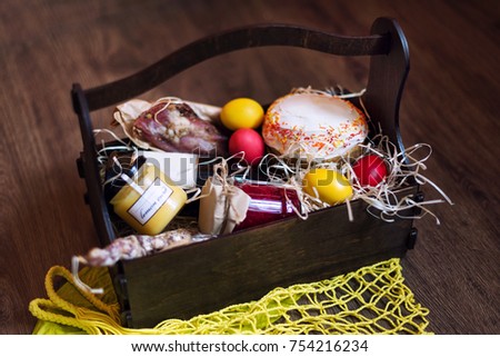 Colorful easter eggs in a basket with cake, red wine, hamon or jerky and dry smoked sausage on wooden background