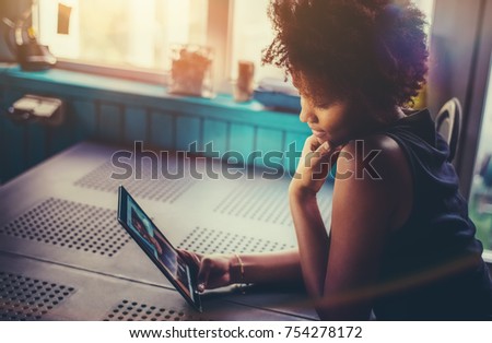 Cute and charming young curly black girl is sitting at the table in dark setting next to window with digital tablet in her hands and browsing just recently taken selfie pictures made on front camera