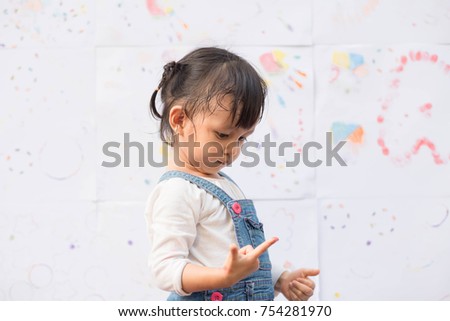  Cute asian little girl is looking her finger covered with the colors in the background of painted and stamp colorful abstract picture, concept of art education and learning by playing for kid.