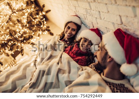 Merry Christmas and Happy New Year! Happy family in Santa Claus hats are lying in bed under the blanket near beautiful Christmas tree. Enjoying being together on New Year's Eve and waiting for holiday