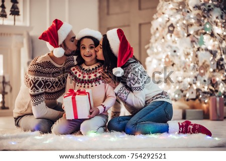 Merry Christmas and Happy New Year! Happy family is sitting near beautiful Christmas tree at home. Parents presenting gift box to their charming daughter and kissing her to cheeks.