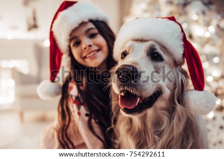 Merry Christmas and Happy New Year! Charming little girl in Santa Claus hat with dog labrador retriever are waiting for the New Year at home, smiling and looking at camera.