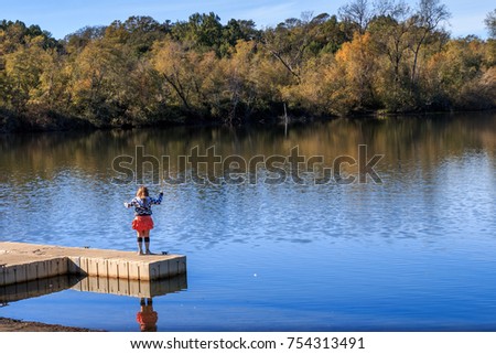 A little girl fishing from a dock on a lake. She has red shorts, black long socks and blue and white jacket. The lake is blue with trees in autumn colors.