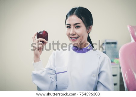 Female dentist holding red apple in her hand while standing in front of dental equipments and devices in clinic. Heath care and maintenance concept with photo filter effect