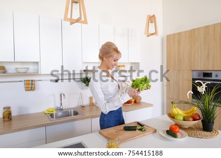Slender girl takes care of figure and prepares light supper, sorts out green salad leaves with smile on face. blonde at top of modern light kitchen with bowl of fruit and vase with potted plant and