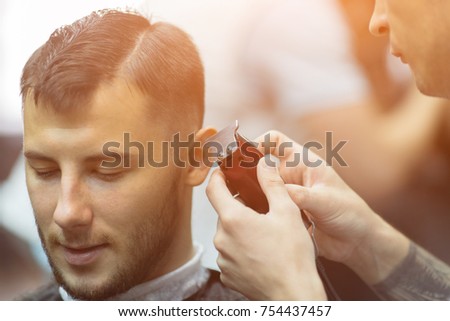 young charming man in barbershop salon while shearing with clipper by hairdresser, sunlight