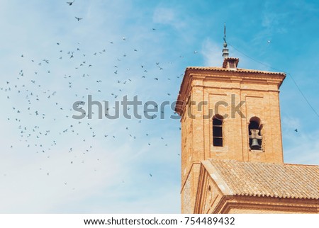 A plenty of pigeons flying over the roof of a bell tower of an ancient catolic church in the blue cloudy sky.