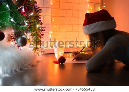 Cute girl writes a letter to Santa Claus near the fireplace and Christmas tree in the light of the garlands