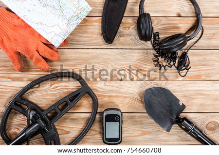 Overhead view of metal detector accessories placed on rustic wooden table. Items included metal detector, shovel, knife, gps, gloves, map and headphones. Treasure hunters concept.