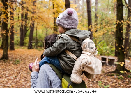 Little girl in autumn forest with teddy bear backpack sitting on mother shoulders. Fall day. Little girl exploring nature with mother.