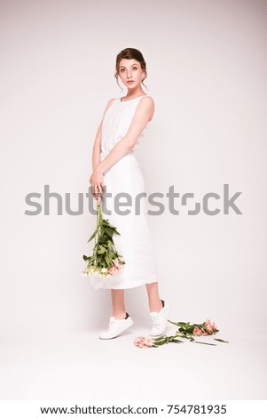 attractive young woman in white dress holding beautiful flowers and looking at camera isolated on grey