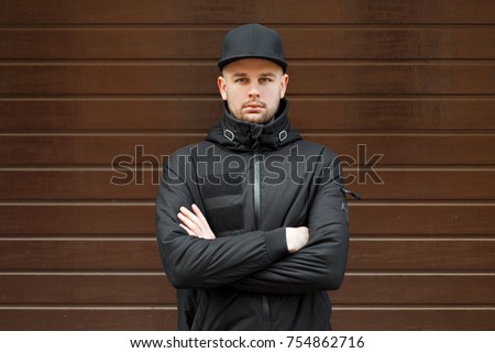 stylish handsome man in a black fashionable baseball cap and black winter jacket stands and looks at the camera near the wooden wall