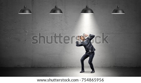 A scared businessman stands cowering under the rays of industrial pendant lights right above him. Shadow business. Found culprit. Caught in the act. Royalty-Free Stock Photo #754865956