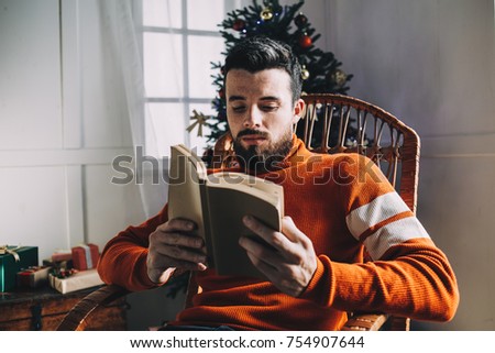Another picture of good-looking guy sitting in the rocking chair at the Christmas tree and reading interesting book. He has almost finished reading it. Man decided to read a book while he is waiting