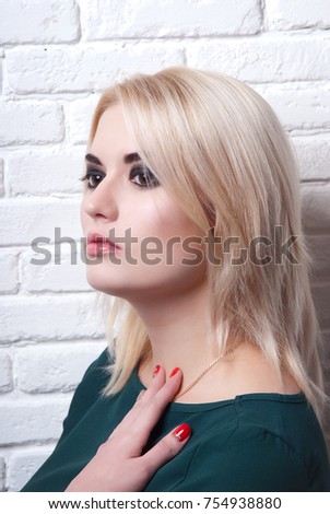 Blonde girl with makeup smoky eyes and a hand on his chest looking to the side in front of a white brick wall