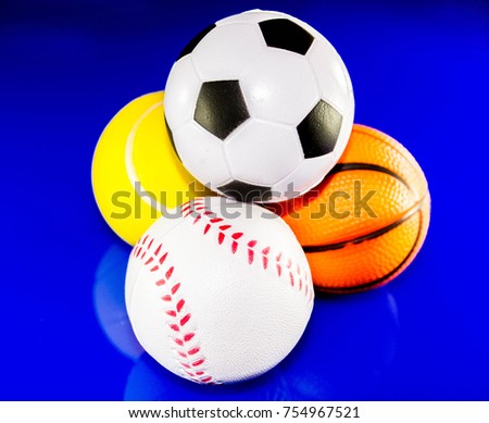A closeup image of a stack of sports balls against a reflective blue background. In the stack is a football, baseball, basketball and tennis ball.