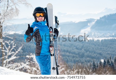 Happy female skier wearing blue ski suit, black helmet and mask smiling showing thumbs up posing in the mountains copyspace