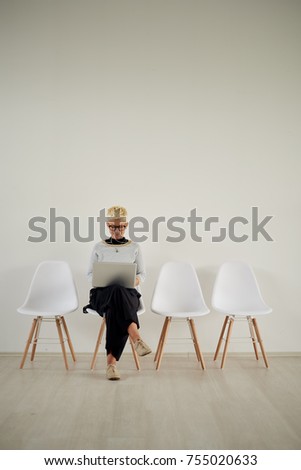 Businesswoman sitting on chair in the lobby and using laptop. White wall in background