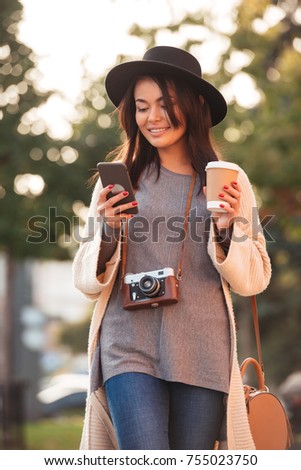 Young attractive asian woman in black hat texting sms on mobile phone and holding coffee cup, while walking in city park