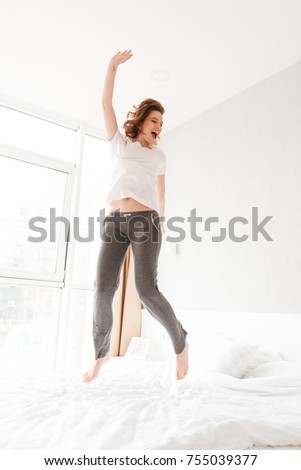 Image of happy amazing young woman in bed indoors jumping. Looking aside.
