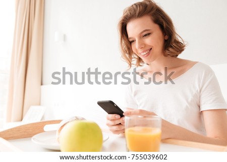 Image of smiling young lady sitting indoors at the table with croissant and juice. Looking aside chatting by phone.