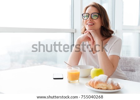 Picture image of cheerful young pretty woman sitting indoors at the table with juice and croissant eating corn flakes. Looking aside.
