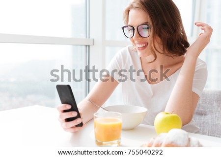 Picture of happy young amazing woman sitting indoors at the table with juice and croissant and corn flakes. Looking aside chatting by mobile phone.