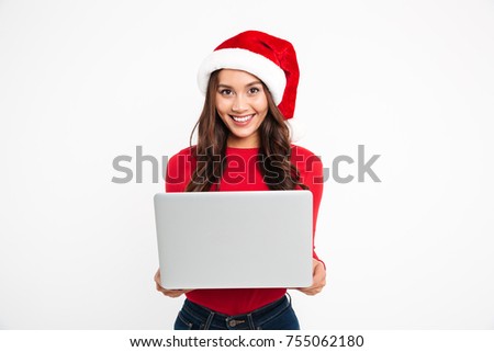 Portrait of a smiling attractive asian girl dressed in red christmas hat holding laptop computer and looking at camera isolated over white background