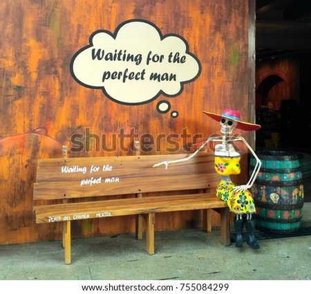Traditional painted Mexican skeleton in yellow dress and pink hat sitting on wooden bench next to funny sign Waiting for the Perfect Man in Playa del Carmen, Mexico – humorous concept of relationship