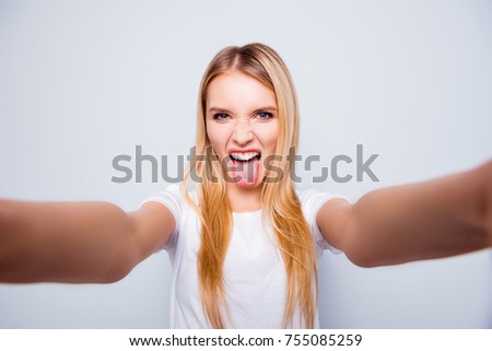 Funky crazy woman with blonde hair is showing her tongue and taking self portrait on her smartphone. She is isolated on grey background