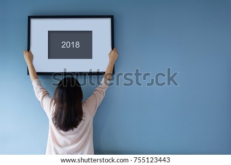Rear view Asian woman holding and and setting picture frame on the blue wall. Female decorating room. Moving and renovate house. 2018, 2020, 2021, Start and change to New Year goals concept