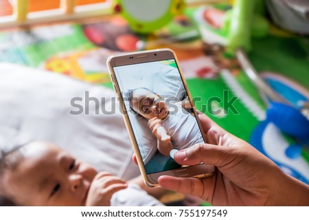 Above high angle shot of happy mom taking photo her Asian baby boy in bed with a smartphone at home. It is a memorable authentic feeling for every parent when their child is growing up. Top view