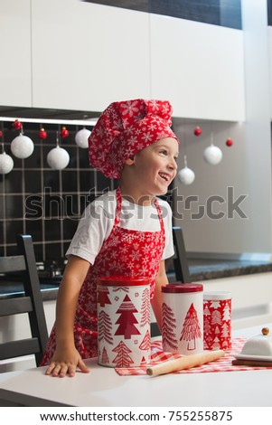 The boy in the Christmas cap of the cook and apron helps her mother cook cookies with ginger in a light kitchen with cans of New Year's pictures.  Happy family, preparing for the holiday
