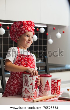 The boy in the Christmas cap of the cook and apron helps her mother cook cookies with ginger in a light kitchen with cans of New Year's pictures.  Happy family, preparing for the holiday