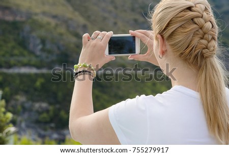 Young beautiful blonde woman doing a picture with your mobile phone. A woman is standing against a background of mountains, she is a tourist. Photograph mobile phone.