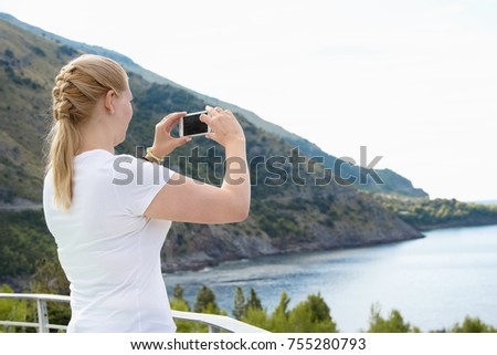 Young beautiful blonde woman doing a picture with your mobile phone. A woman is standing against a background of sea and mountains, she is a tourist. Photograph 