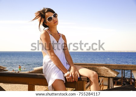 Young beautiful girl sits on the beach and smile. Beautiful teenager dressed in white dress and sunglasses sitting on the fence, in the background is the blue sea and sky
