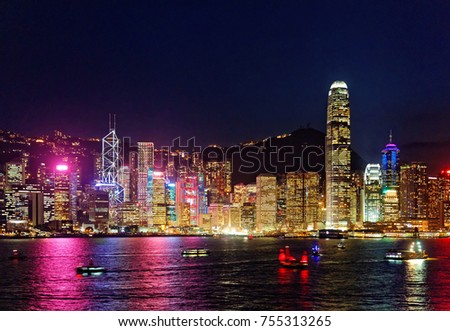 Majestic urban skyline of crowded skyscrapers by Victoria Harbour in Hong Kong at night, with colorful city lights glistening & reflected in the water and ferry boats crossing the seaport in the dark