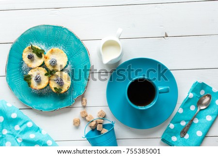 cheesecakes decorated by  frozen  blackberries and mint,  tea in blue cup, milk jug and cane sugar in baby bucket, blue napkin at white polka dots on white wooden table, top view