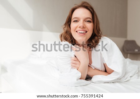 Image of young cheerful lady lies on bed indoors home. Looking camera.