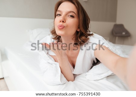 Image of young lady lies on bed indoors home. Looking camera blowing kisses to you.