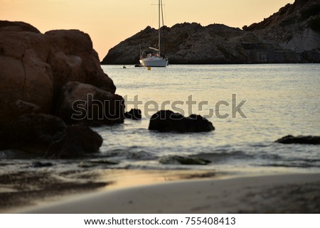 Stunning picture of a sunset at the beach, in Cala Tarida at Ibiza. Focused delicated boat at the background of the photography and kind of blurred stones and shore at the foreground. Contrasted.