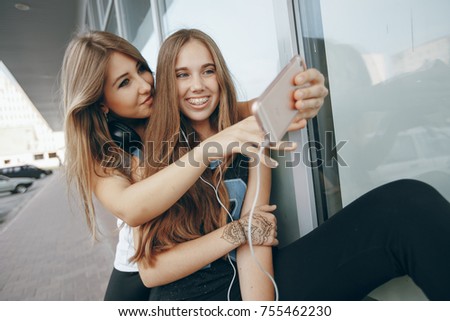 Two beautiful girls at the window wall with headphones taking pictures on a phone
