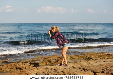 A young blonde woman wearing short shorts and plaid shirt taking pictures on a beach. 
Standing on sea rocks; sea, sky, clouds in background. 